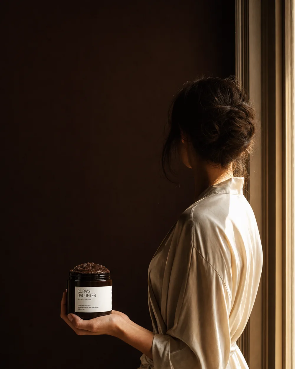 Bride in a cream robe at a window holding a jar of CoBa’s Daughter Robusta coffee scrub