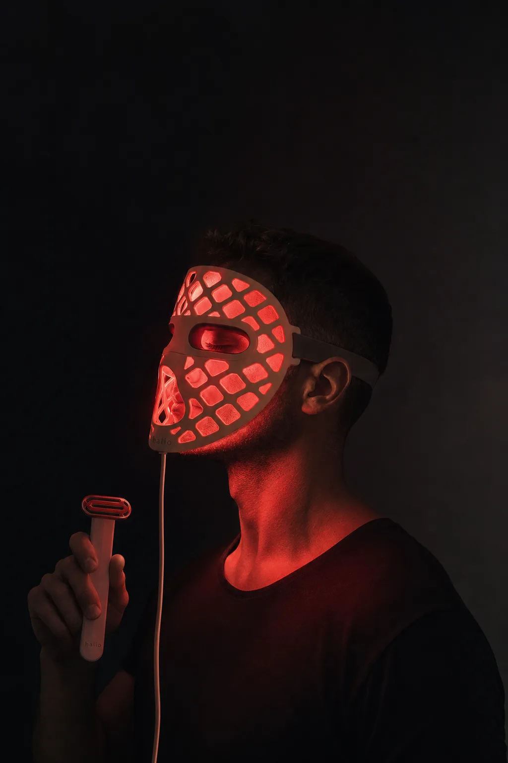 Man in side profile wearing the lit PureGlow mask, holding the red light wand, in near darkness